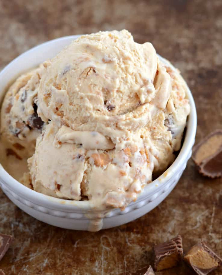 Scoops of peanut butter chocolate ice cream in a small white bowl, with crushed chocolate candy bars scattered around on a rustic brown surface.