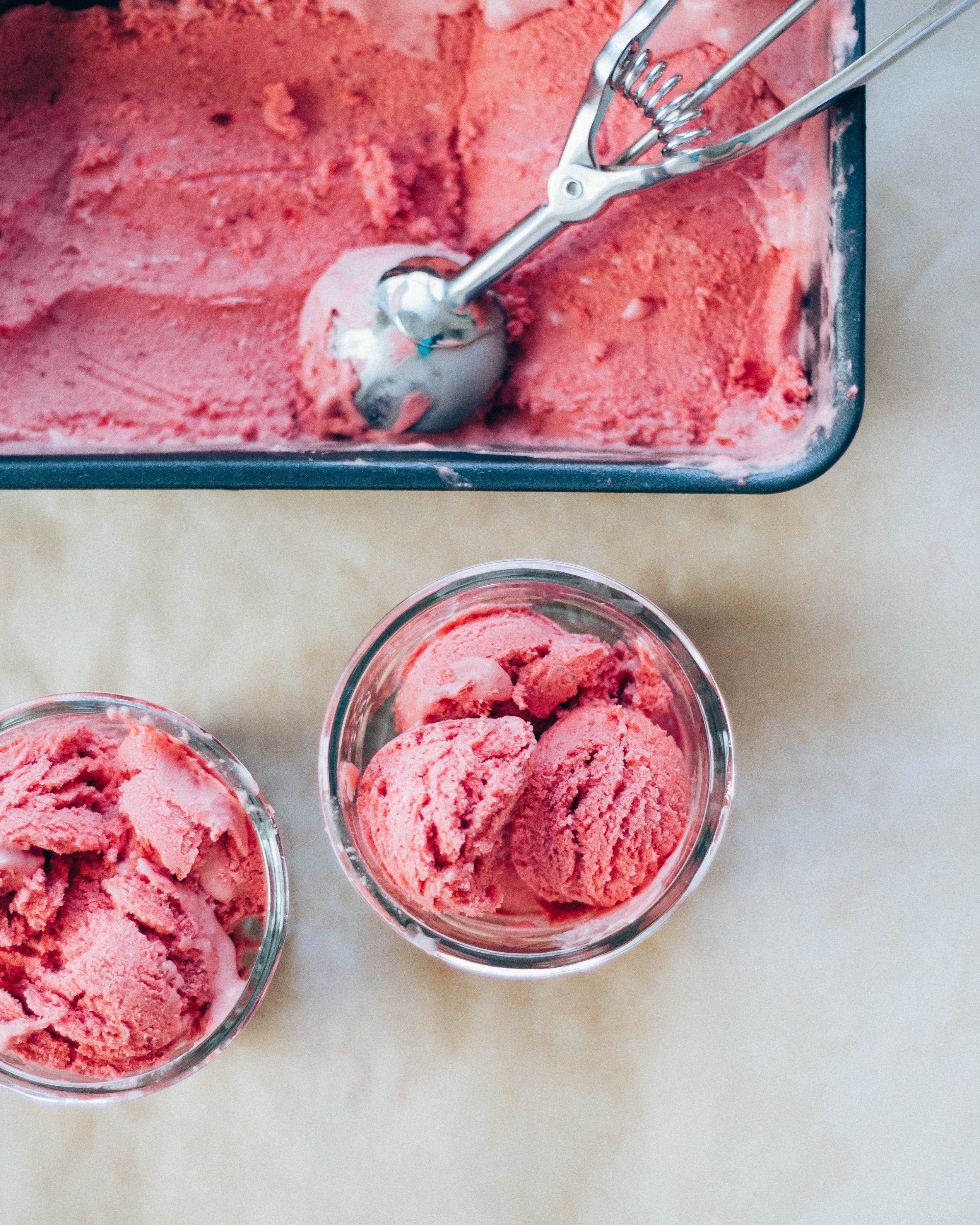 Pink ice cream in a container with two scoops in small bowls on a light background